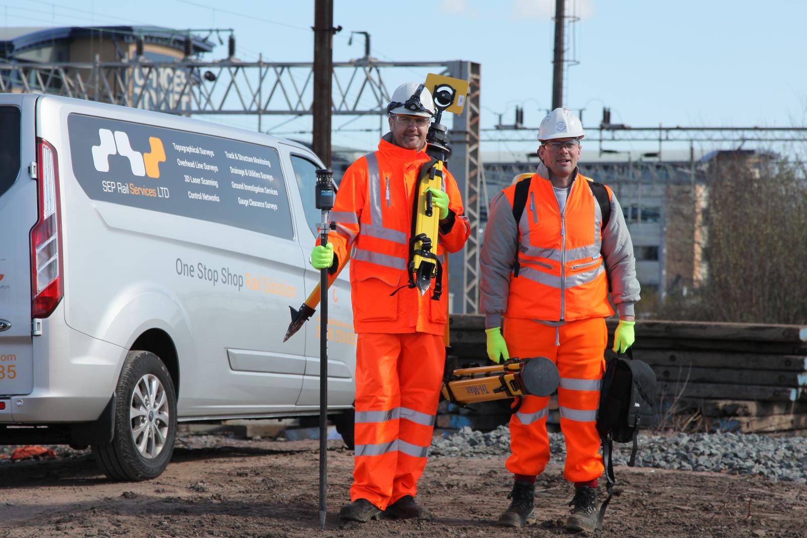 Two men stood holding equipment on a railtrack, in front of a van