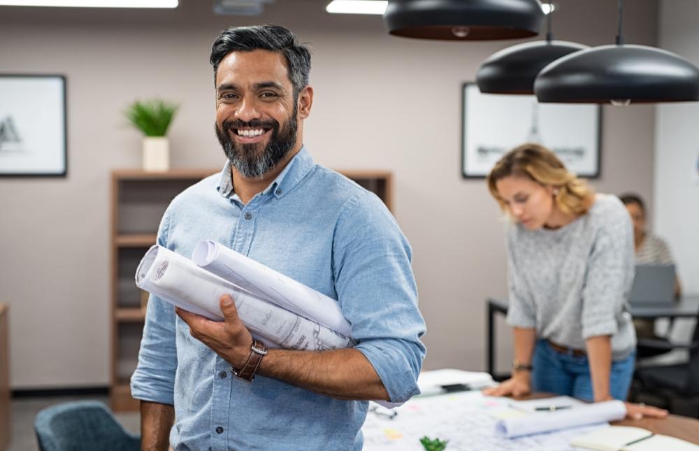 Man in a blueshirt holding rolls of paper, in front of a woman stood at a desk