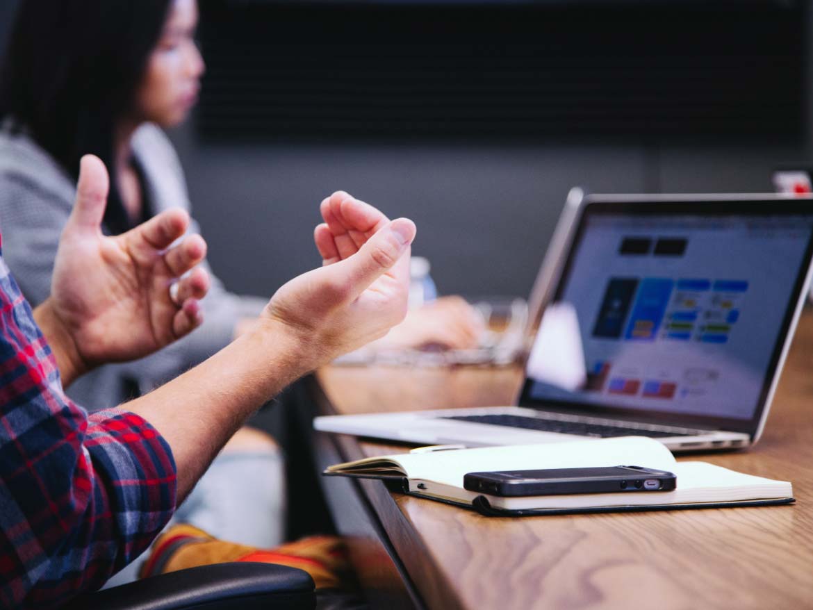 Arms gesturing in front of a white laptop with a woman in the background