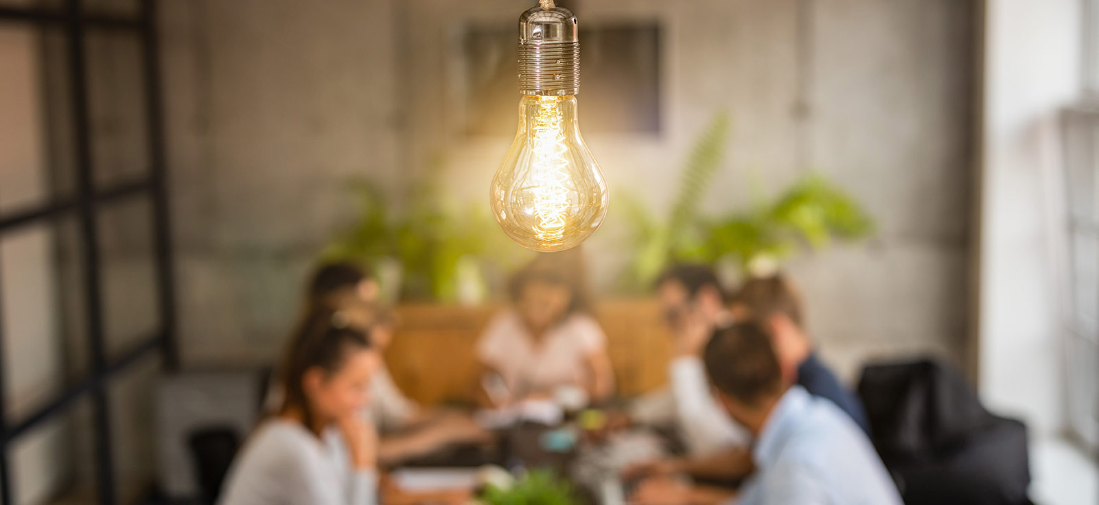 Yellow lit lightbulb in front of a blurred background of 6 people sat around a table 