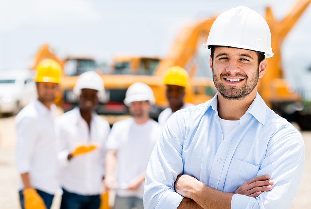 A man with his arms folded wearing a white construction hat with workers in the background