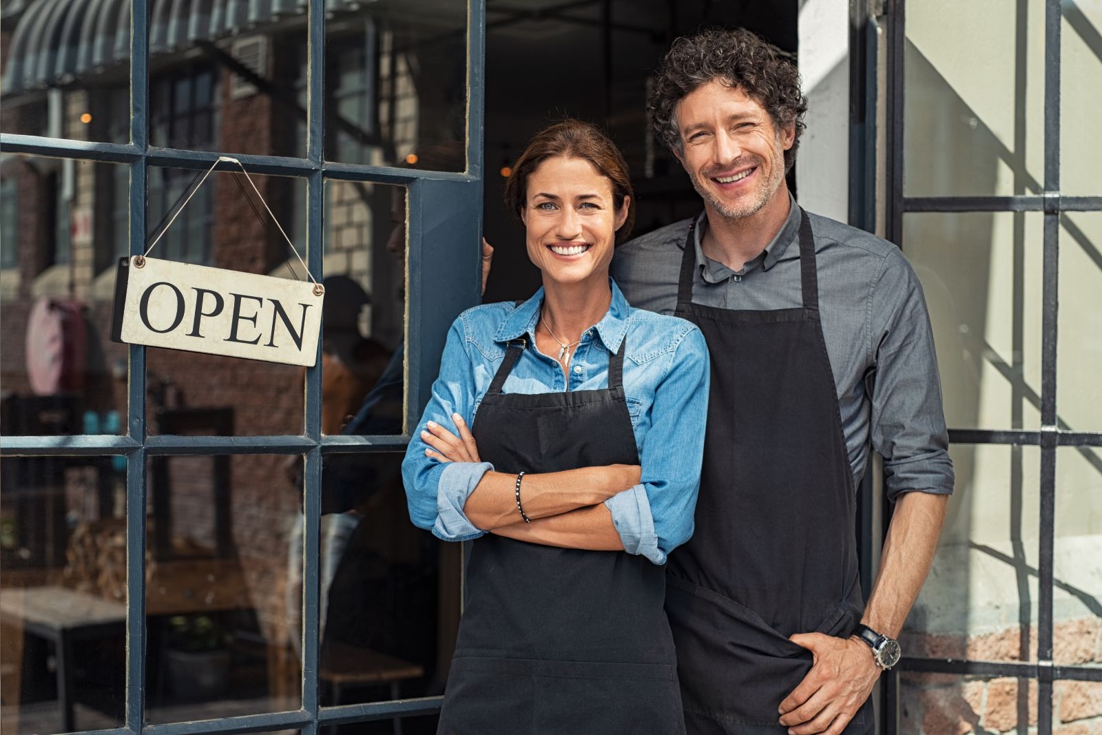 Man and a woman wearing aprons stood in front of an shop with an open sign