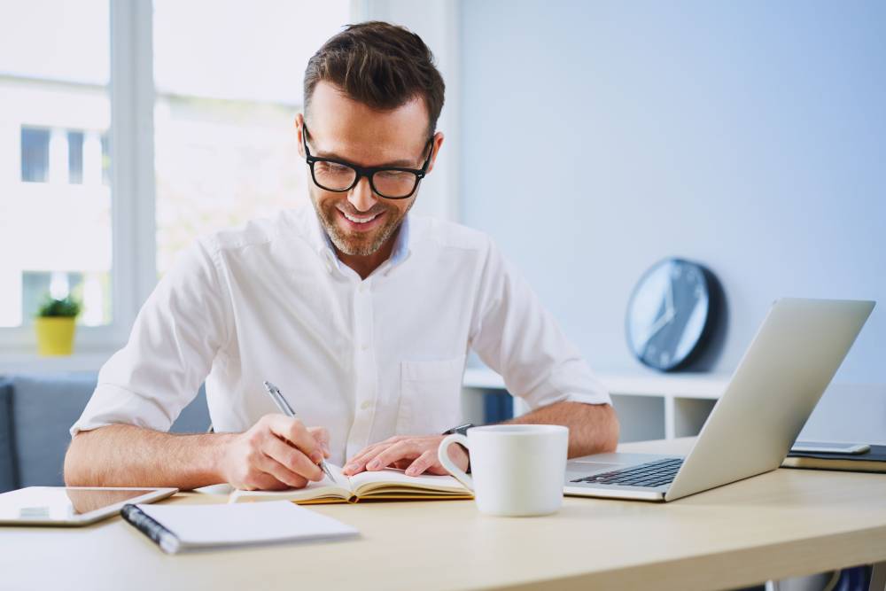 Man wearing glasses writing at a desk in behind a laptop