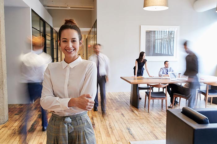 Woman standing smiling in a busy office space as people move around her